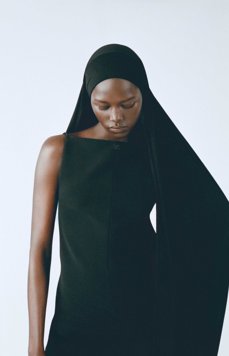 Elegant dark-skinned woman in black dress and head covering, eyes downcast, against a clean white backdrop in a studio fashion shot.