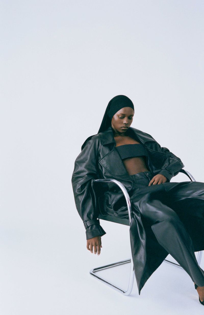 An adult woman in a black leather outfit is sitting on a metal chair against a white background in a fashion photography studio.