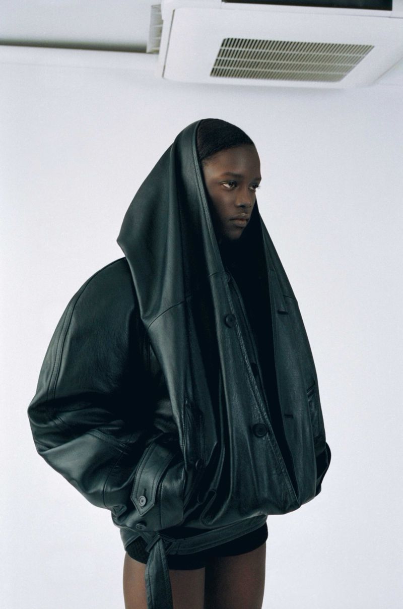 A person in a hooded black leather jacket stands in front of a white wall. The jacket is oversized and the setting is a minimalist photo studio.