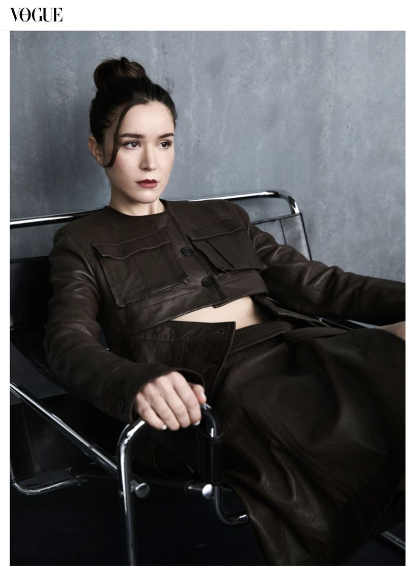 Fashion editorial shot of a woman with her hair in a bun, wearing a dark brown leather jacket and skirt, sitting in a chrome chair.