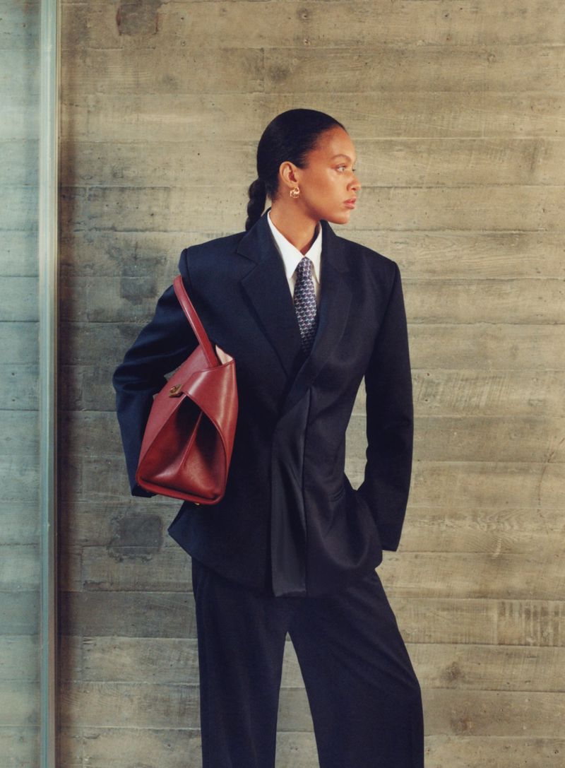 Model in a dark oversized suit carrying a red leather handbag against a concrete wall for Vogue Espana.