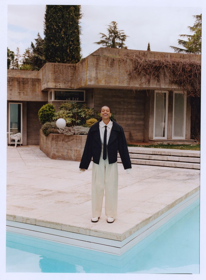 Stylish woman in oversized jacket and wide-leg pants poses by a pool next to a brutalist concrete house with lush greenery.