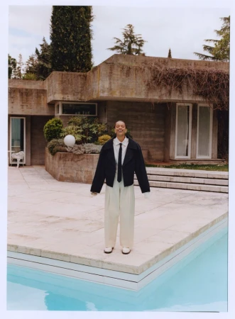 Stylish woman in oversized jacket and wide-leg pants poses by a pool next to a brutalist concrete house with lush greenery.