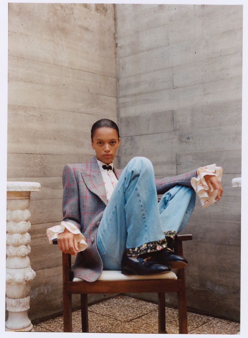 Woman in tweed blazer and jeans sits on a wooden chair against a textured wall in a fashion editorial shot with neutral light.