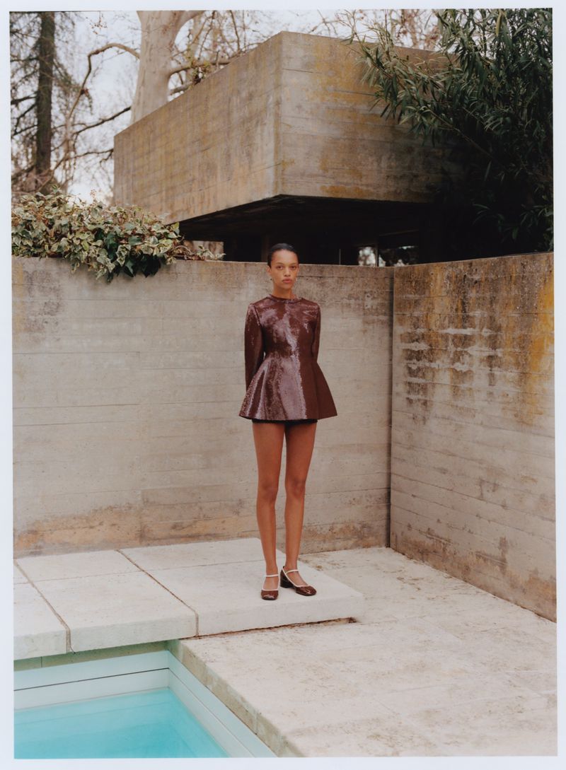 Fashion shot of an adult woman in a brown dress posing next to a pool with a modern, concrete building in the background.