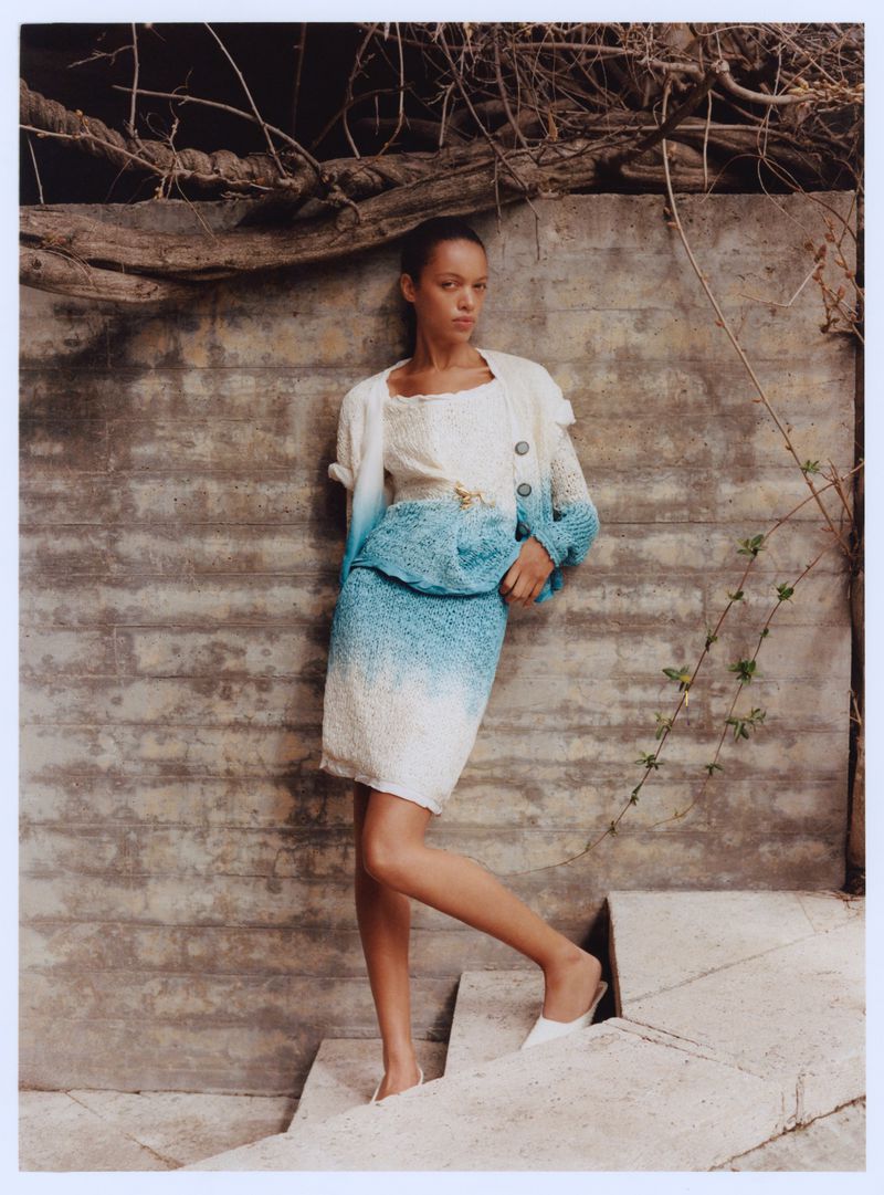 A woman in a blue and white skirt and sweater poses on concrete stairs next to a textured wall with vines in natural light.