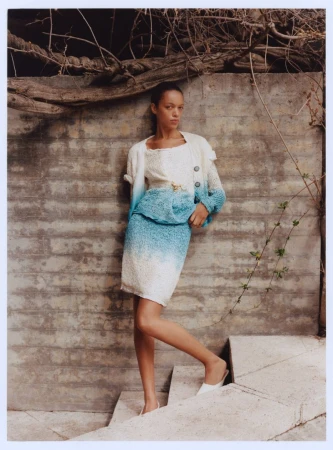 A woman in a blue and white skirt and sweater poses on concrete stairs next to a textured wall with vines in natural light.