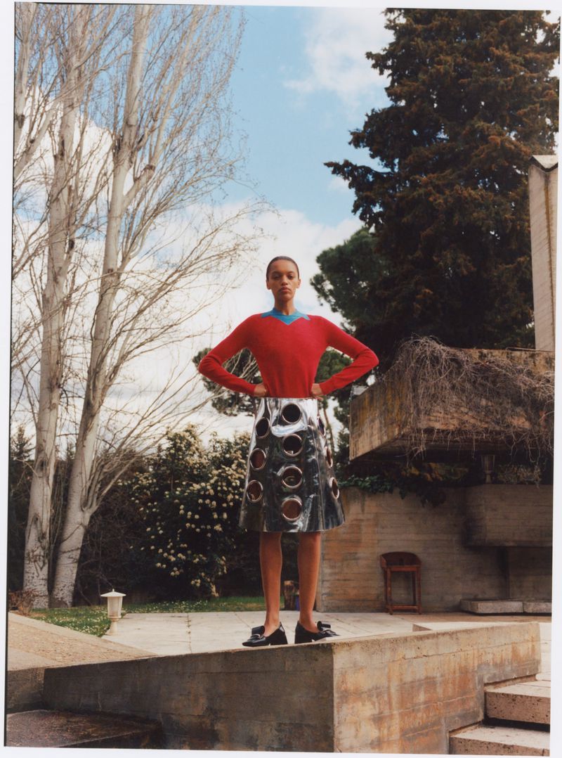 Fashion editorial shot of an adult woman standing outside wearing a red sweater and skirt on a concrete surface with trees and sky visible.