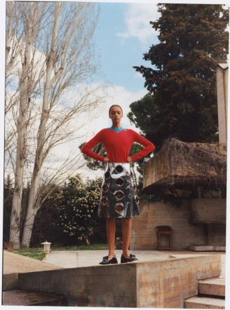 Fashion editorial shot of an adult woman standing outside wearing a red sweater and skirt on a concrete surface with trees and sky visible.