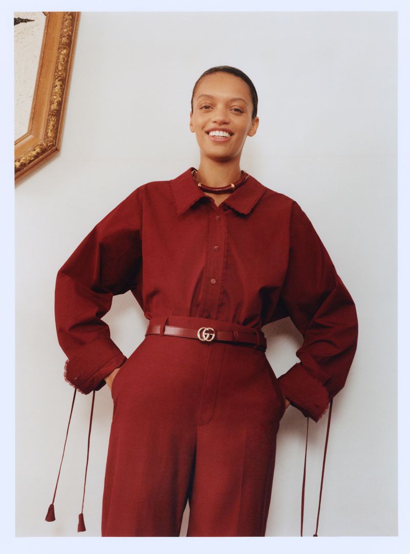 A fashionable Black woman smiles in a maroon shirt and pants, cinched with a logo belt, inside a bright room with a classic framed picture.