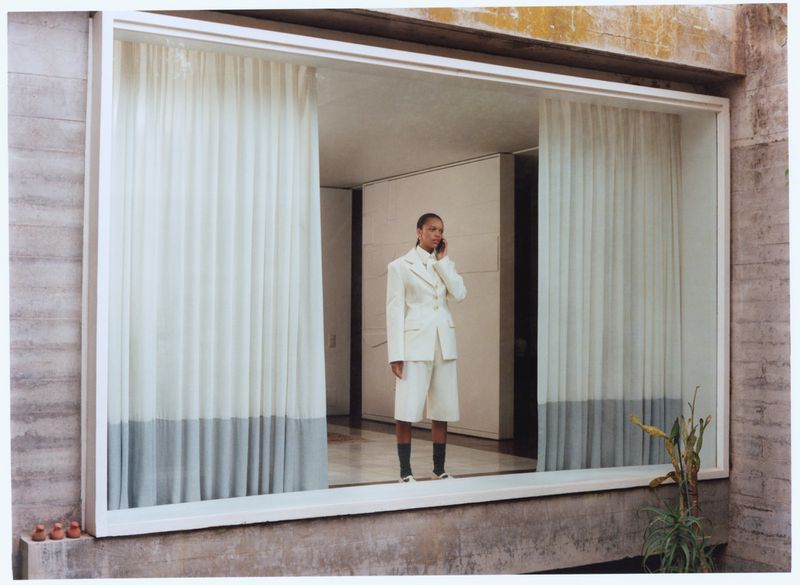 An adult woman wearing a white suit and black socks talks on a cell phone, standing behind a framed window with sheer white curtains.