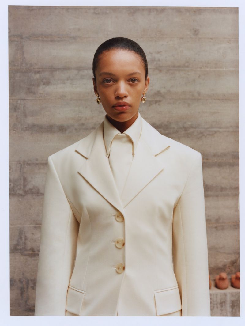 Woman in a white suit stands indoors against a textured wall, gazing directly at the camera in a fashion portrait with neutral lighting.