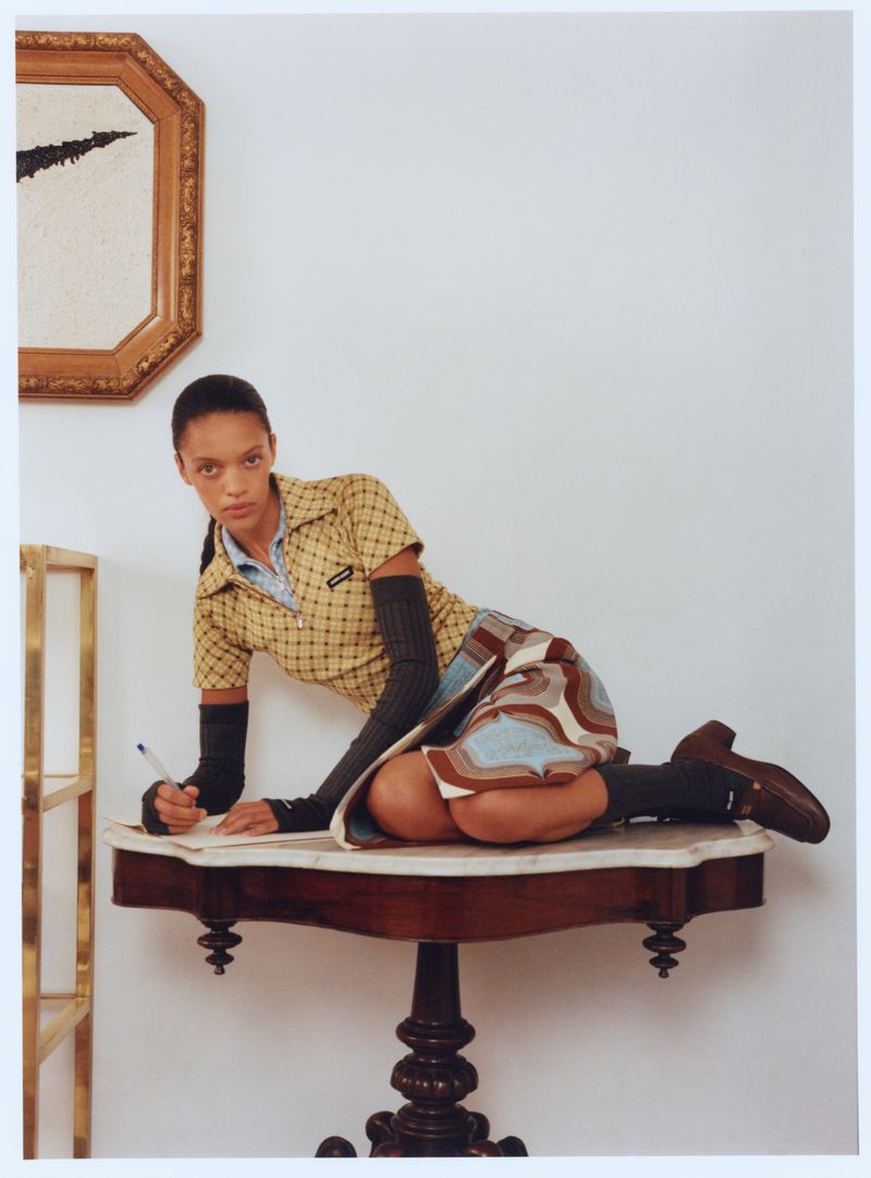 A woman in a patterned yellow shirt and patterned skirt poses on a brown marble table next to a white wall with framed art.