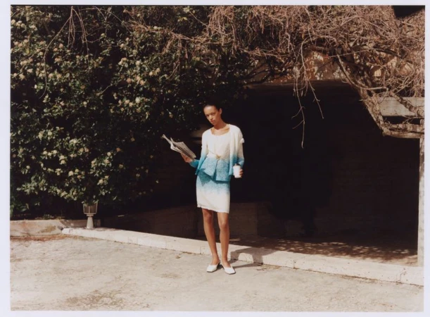 Woman in a skirt suit reading a newspaper in front of foliage and a building exterior on a bright day with a coffee cup.