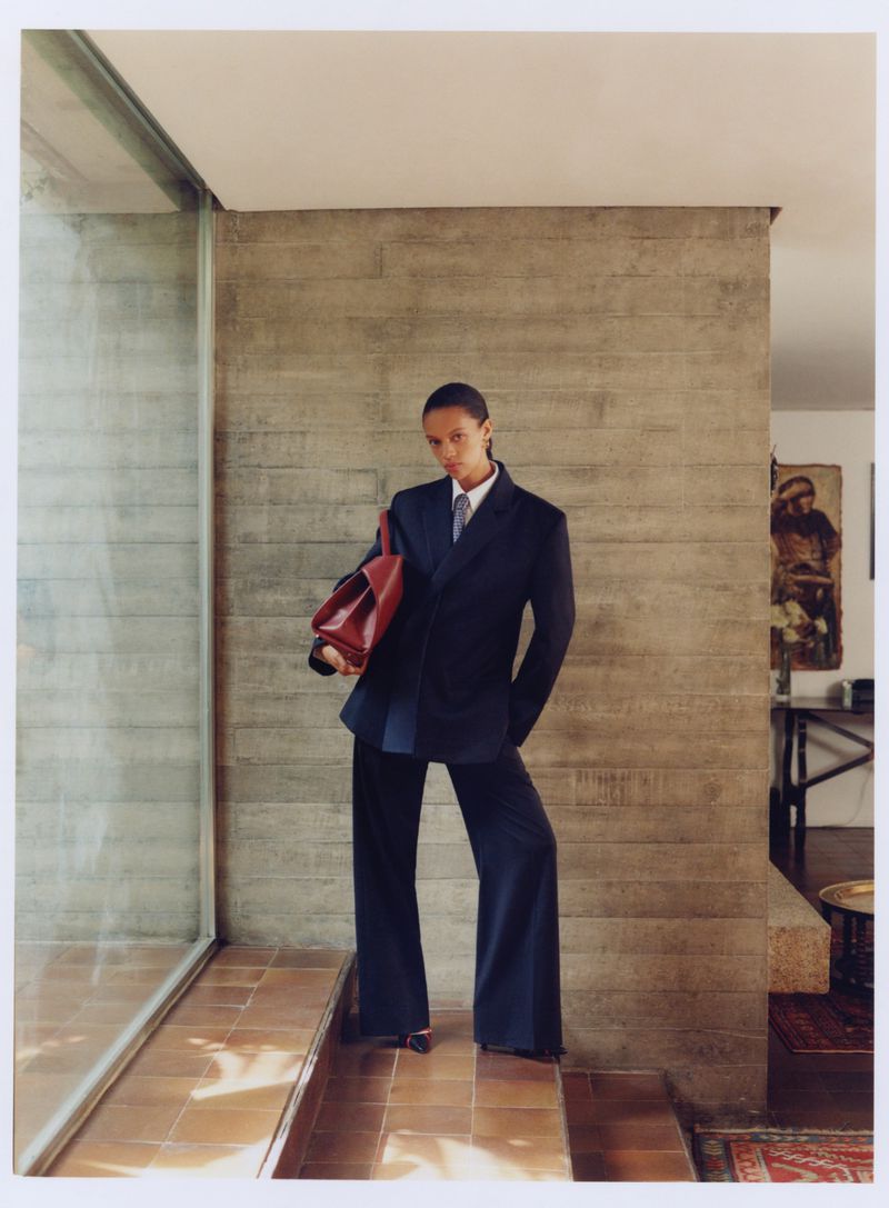 Fashion shot of a woman in a blue blazer and pants holding a red bag. Indoor setting with a modern wall and tiled floor.