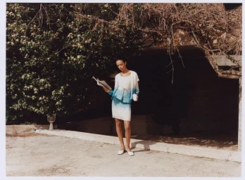 Model standing outdoors holding a newspaper and a cup against a stone wall and flowering bush, wearing a blue and white knit skirt suit.