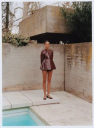 Model standing outdoors near a concrete architectural structure and swimming pool wearing a dark glossy peplum top.