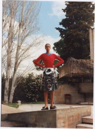 Model standing outdoors wearing a red top and metallic cutout skirt against a stone terrace and trees.