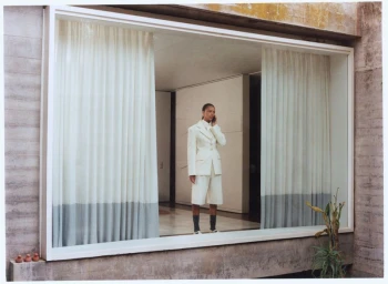 Model standing behind a large architectural window framing a minimalist interior with white curtains.