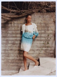 Model leaning against a weathered stone wall wearing a blue and white ombre knit cardigan and skirt set on outdoor steps.