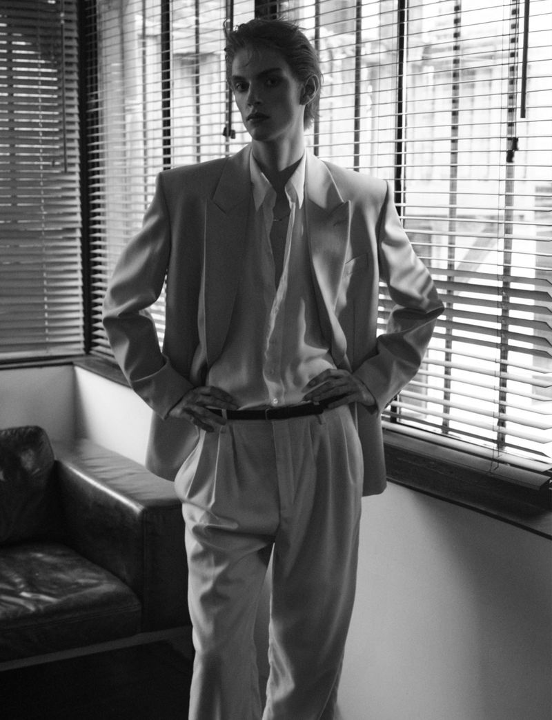 Black and white portrait of an androgynous young adult in a suit, standing near window blinds in a studio setting. Fashion editorial.