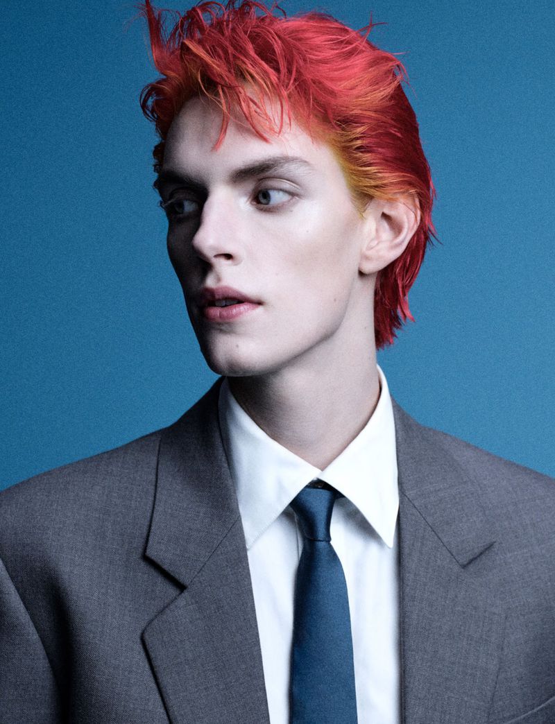 Portrait of a person with red and blonde dyed hair wearing a grey suit, white shirt and blue tie against a blue background.