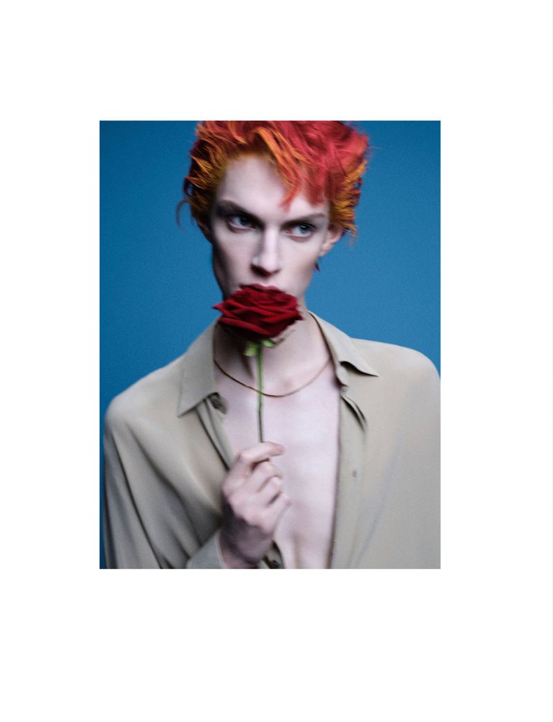 Close-up portrait of a model with fiery red and orange hair holding a red rose to their lips against a solid blue backdrop.