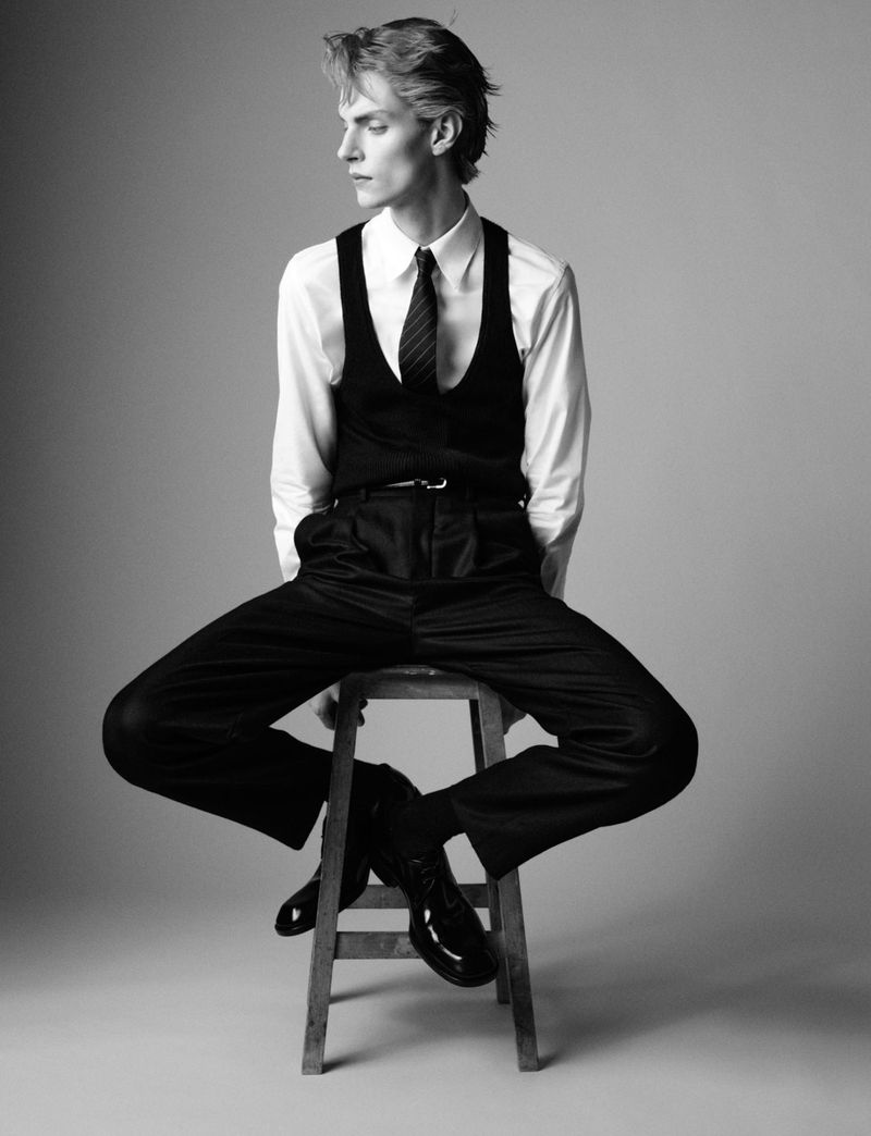 Monochrome fashion portrait of a male model wearing a white shirt, black vest, and dark trousers seated on a wooden stool.