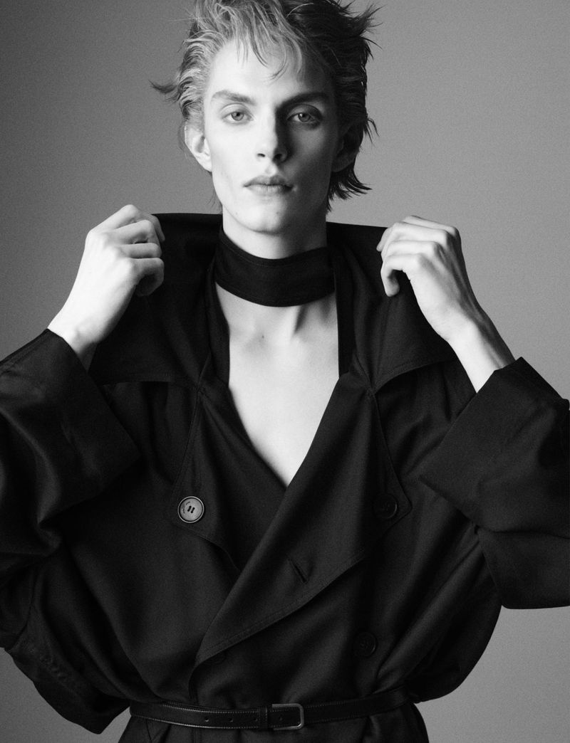 Black and white portrait of a young adult man with slicked-back blonde hair, wearing a dark trench coat and neck scarf against a grey backdrop.