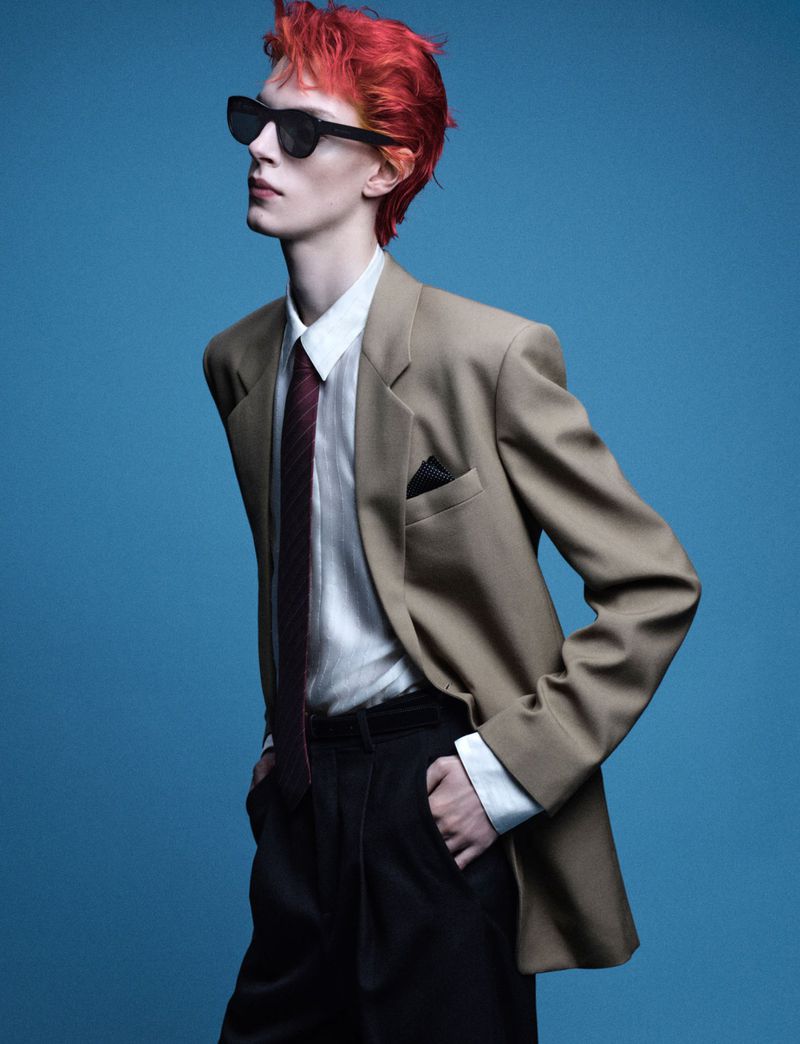 Adult male with dyed red hair and sunglasses poses in a tan suit jacket, white shirt, and black tie against a blue background.