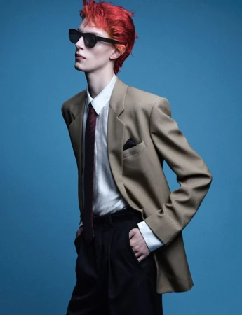 Adult male with dyed red hair and sunglasses poses in a tan suit jacket, white shirt, and black tie against a blue background.