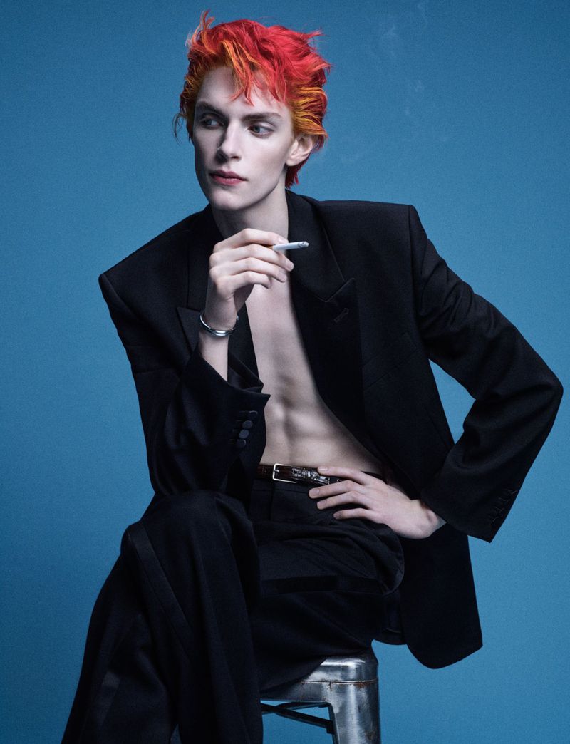 Model with bright red and orange hair holding a cigarette while wearing a black blazer against a blue background