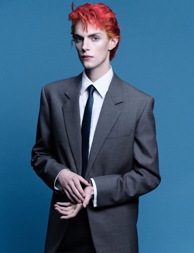 Androgynous male model with striking orange hair, wearing an oversized grey suit and tie against a solid blue background.