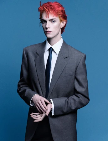Androgynous male model with striking orange hair, wearing an oversized grey suit and tie against a solid blue background.