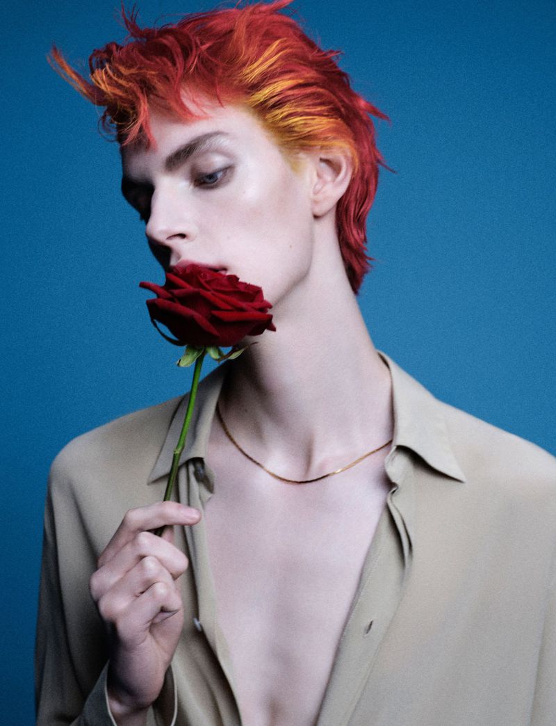 Androgynous person with red and yellow hair holds a red rose against a blue background. Portrait-style shot with neutral clothing.