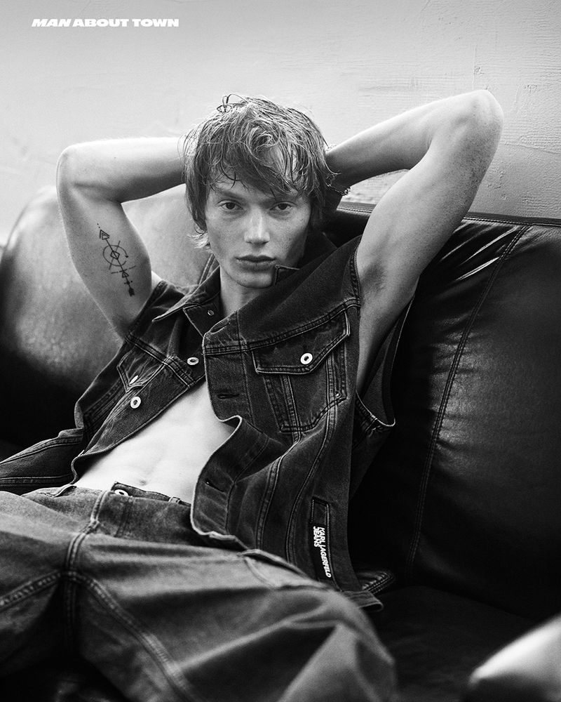 Black and white studio shot of a young adult male with dark hair, denim jacket and pants, posing on a black leather sofa with his hands behind his head.