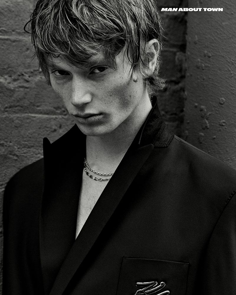 Monochrome portrait of a young man wearing a black jacket and chain necklace standing against a brick wall in an urban environment.
