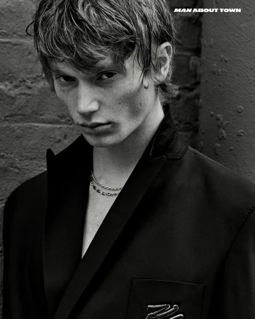 Monochrome portrait of a young man wearing a black jacket and chain necklace standing against a brick wall in an urban environment.