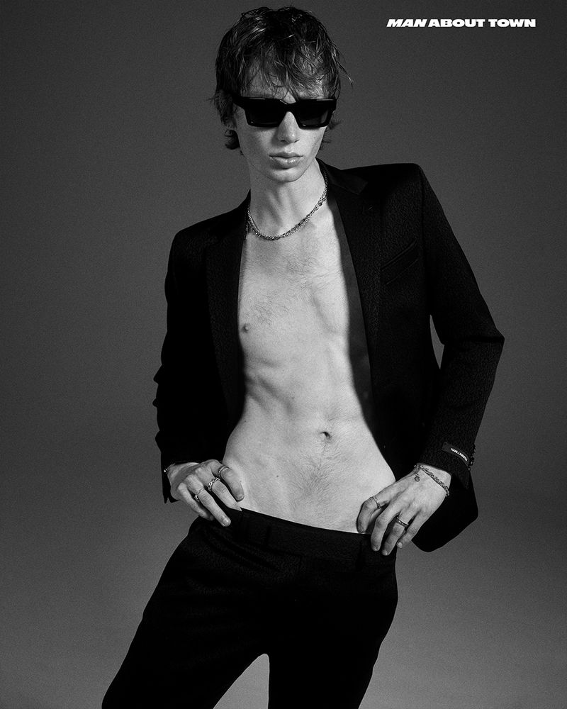 Black and white portrait of a slender man with sunglasses, open blazer, and dark pants posing in a studio setting with a serious expression.