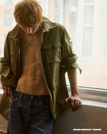 Calum Harper wearing an open olive green jacket and blue denim jeans standing indoors near a large window for Man About Town.