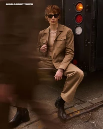 Calum Harper wearing a tan jacket and trousers seated by a vehicle for Man About Town editorial