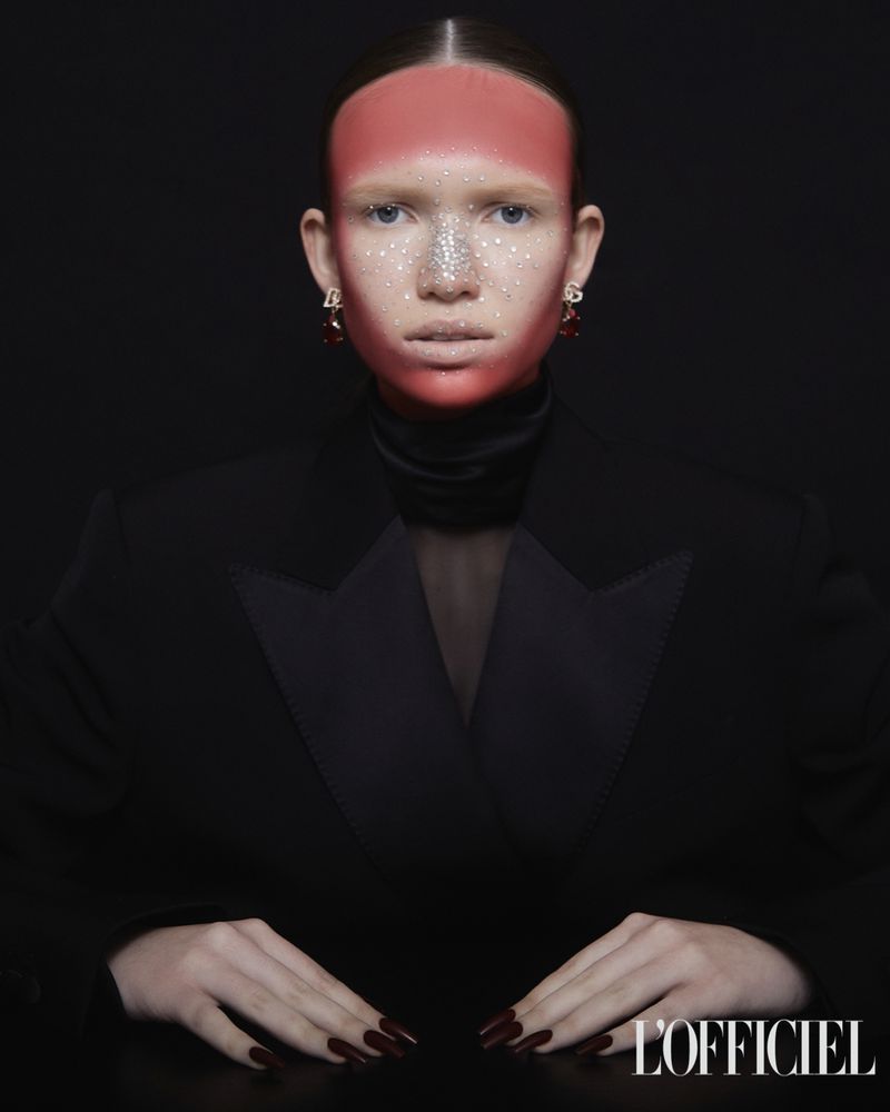 Fashion shot of a woman wearing a black blazer with a red and silver face makeup design, jewelry earrings, and red painted fingernails.