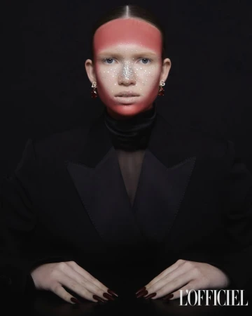Fashion shot of a woman wearing a black blazer with a red and silver face makeup design, jewelry earrings, and red painted fingernails.