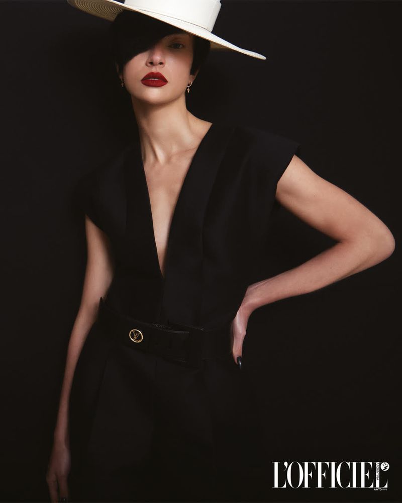 A stylish woman poses against a black backdrop wearing a black low-cut dress, white sunhat, and bold red lipstick in a studio fashion portrait.