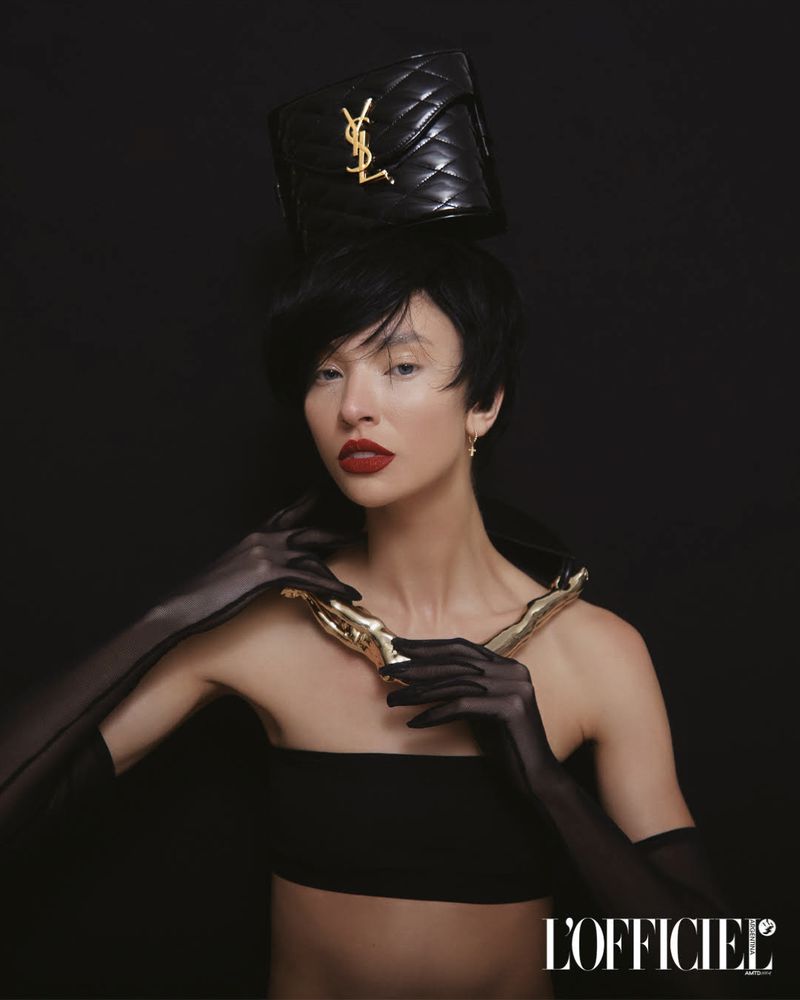 Woman in a black top poses with a gold necklace and a black YSL bag on her head in a studio with a black background.