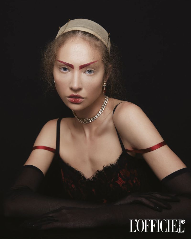 Young woman with dramatic red eyebrows, wearing a black and red lace dress and jeweled necklace, posing against a dark backdrop.