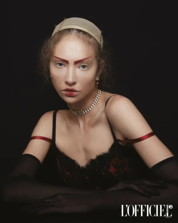 Young woman with dramatic red eyebrows, wearing a black and red lace dress and jeweled necklace, posing against a dark backdrop.