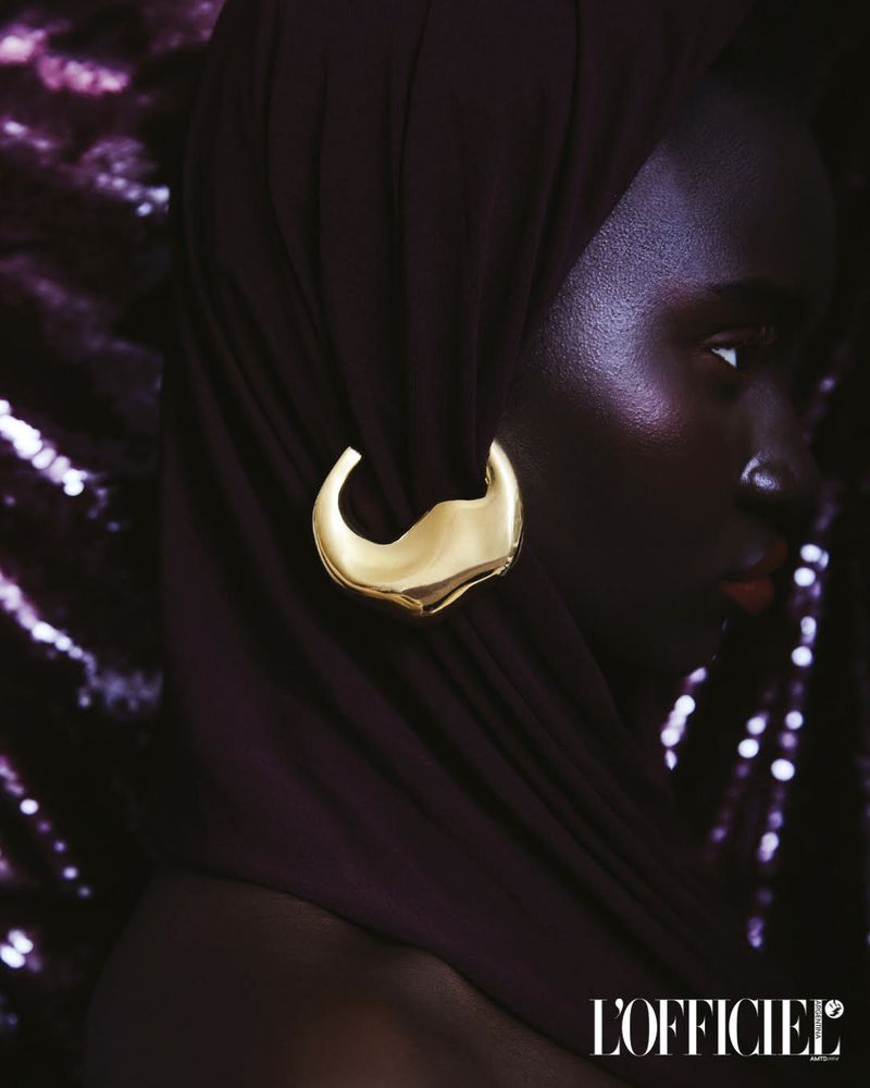 Close-up studio shot of an adult Black woman with golden earring and dark head covering, against a shimmering purple background.