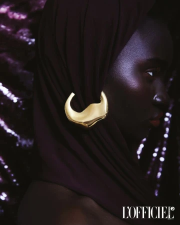 Close-up studio shot of an adult Black woman with golden earring and dark head covering, against a shimmering purple background.