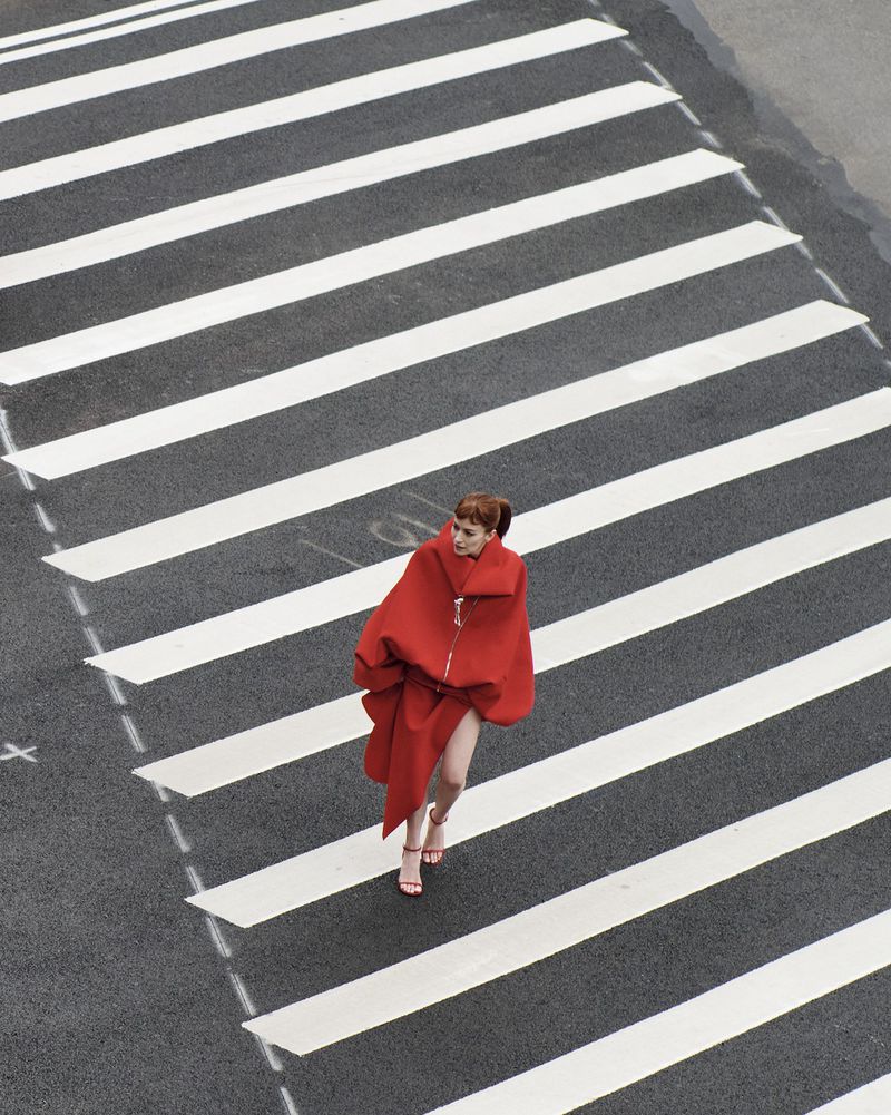 Aerial shot of a woman with red hair and a striking red outfit walking across a black and white striped crosswalk in an urban setting.